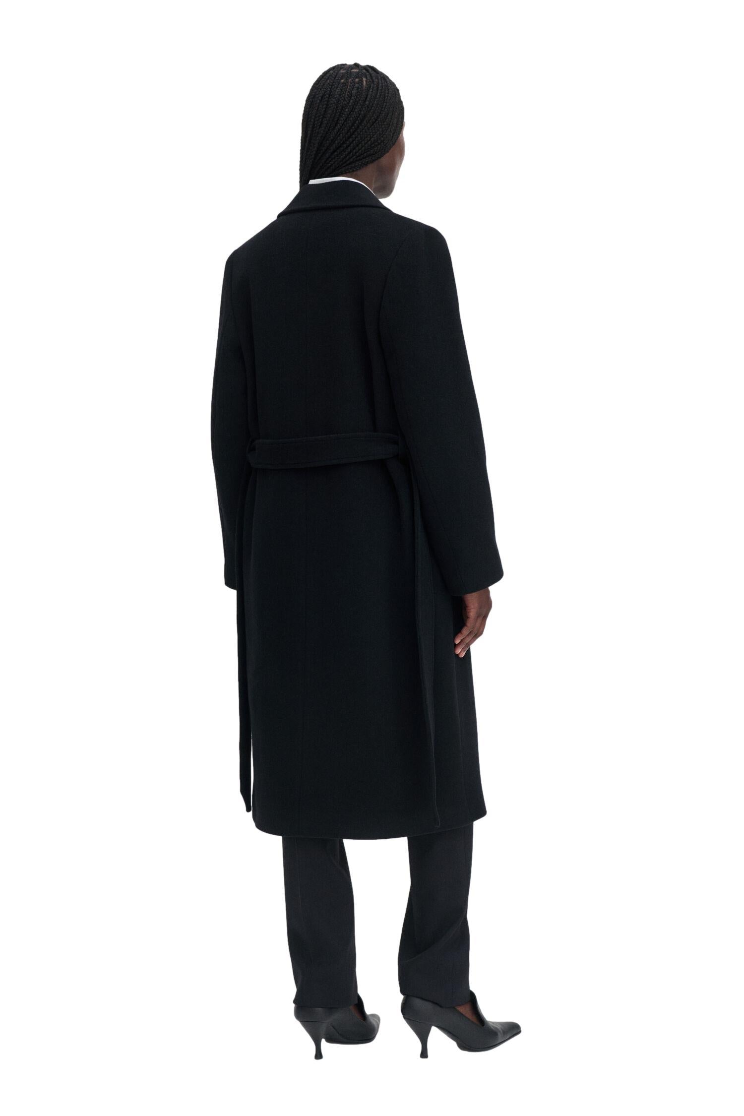 Sort Filippa K Charlotte ullkåpe med belte, lang modell, vises med krage og knapper foran.